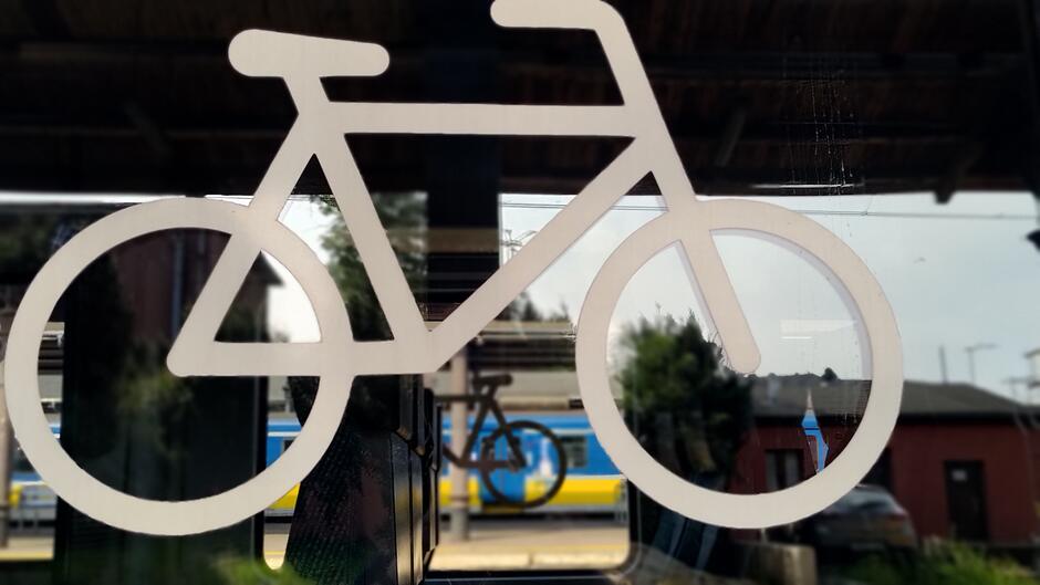 Zdjęcie przedstawia białą ikonę roweru na szklanej powierzchni, za którą widać stację kolejową oraz stojący pociąg w niebiesko-żółtych barwach. W tle znajdują się budynki oraz fragment peronu, co sugeruje, że zdjęcie zostało wykonane na stacji kolejowej.
