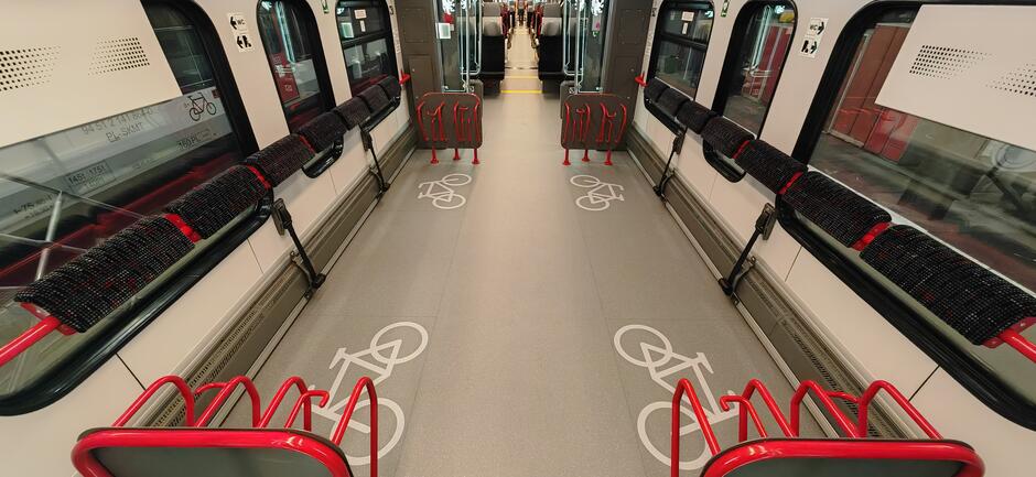 Zdjęcie przedstawia wnętrze wagonu kolejowego, które jest przystosowane do przewozu rowerów. Na podłodze znajdują się oznaczenia rowerów, a po obu stronach wagonu zainstalowane są czerwone uchwyty do mocowania rowerów, nad którymi umieszczone są składane siedzenia dla pasażerów.