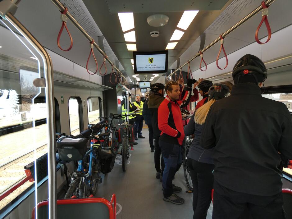 Zdjęcie przedstawia wnętrze wagonu kolejowego, w którym znajduje się grupa pasażerów z rowerami. Pasażerowie, ubrani w odzież sportową i kaski rowerowe, rozmawiają ze sobą, a rowery są umieszczone w specjalnych uchwytach na podłodze wagonu.