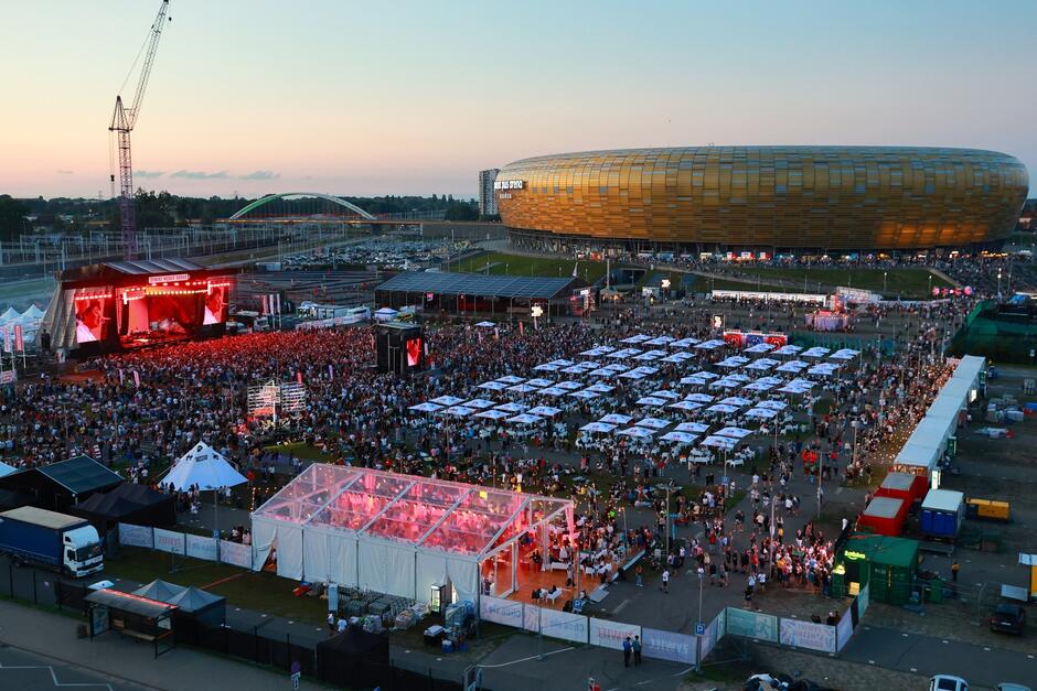 Zrobione z oddalenia zdjęcie terenu koncertu z publicznością i występem na scenie. W tle stadion Polsat plus Arena Gdańsk