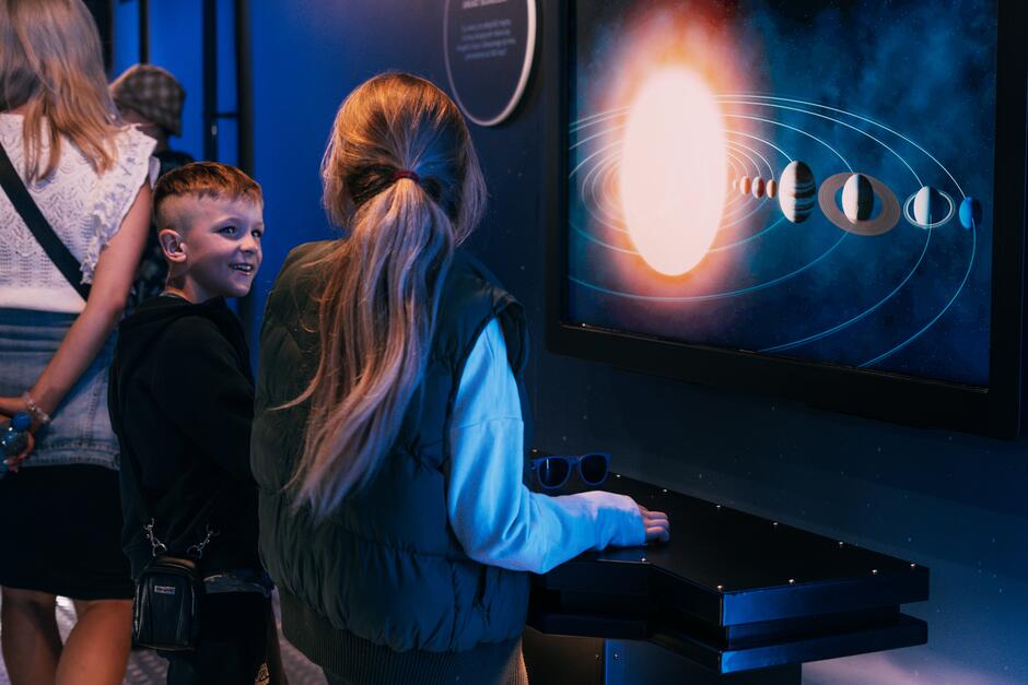 Na zdjęciu dwoje dzieci ogląda ekran przedstawiający schemat Układu Słonecznego z Słońcem i planetami. W tle widać niebieskie oświetlenie wystawy oraz innych zwiedzających.