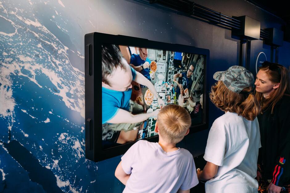 Na zdjęciu troje dzieci ogląda ekran pokazujący astronautów jedzących w kosmosie, prawdopodobnie na Międzynarodowej Stacji Kosmicznej (ISS). W tle widać ścianę z grafiką przedstawiającą widok na Ziemię z kosmosu.