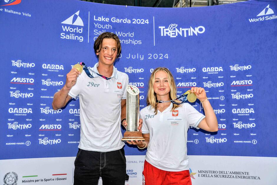 Wysoki młody mężczyzna i niższa młoda kobieta stoją z medalami w dłoniach. Uśmiechają się, w tle ścianka z logo różnych firm