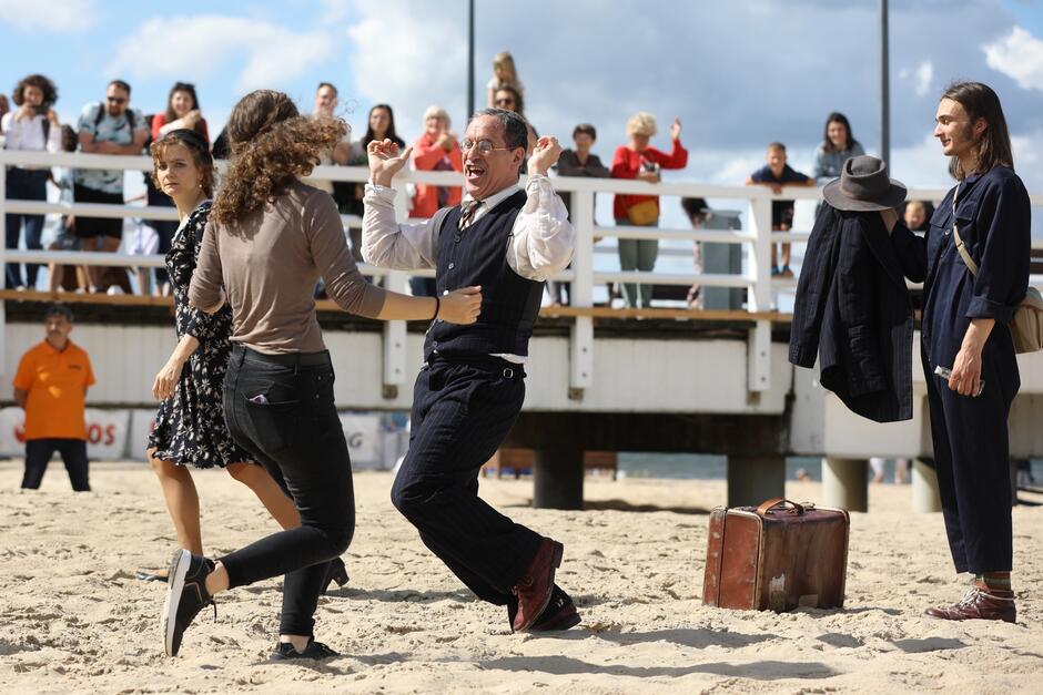 Czwórka aktorów występuje na plaży, obserwują ich widzowie, stojący na molo. Dwoje aktorów - kobieta i mężczyzna, tańczą, obok nich stoi walizka. Obok przechodzi kobieta, spoglądając na tańczących. Inny mężczyzna stoi obok i trzyma na ręku marynarkę oraz kapelusz.