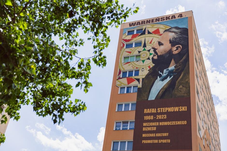 Na zdjęciu widać mural przedstawiający Rafała Stepnowskiego na ścianie wysokiego budynku przy ulicy Warneńska 4. Mural zawiera również napisy: Rafał Stepnowski 1968-2023, wizjoner nowoczesnego biznesu, mecenas kultury, promotor sportu 