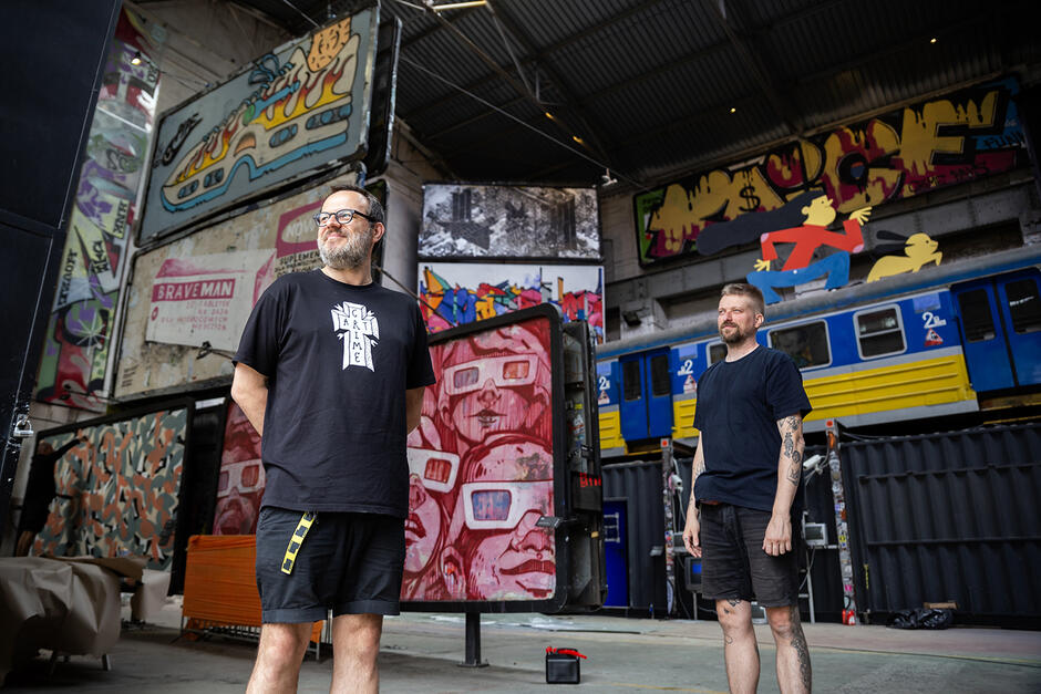 Dwóch mężczyzn stoi w otwartej bramie dawnej hali stoczniowej zbudowanej z cegły, zamienionej na galerię sztuki ulicznej. W hali widać malunki wykonane różnymi technikami na murach oraz różnych nośnikach nośnikach reklamowych, jak billboardy.