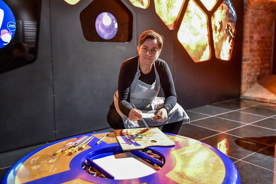 Na zdjęciu widzimy kobietę malującą przy okrągłym stole, na którym znajdują się różne narzędzia artystyczne, takie jak pędzle i kredki. Tło składa się z ciemnej ściany z podświetlonymi elementami w kształcie sześciokątów, które dodają artystycznego klimatu całej scenie.