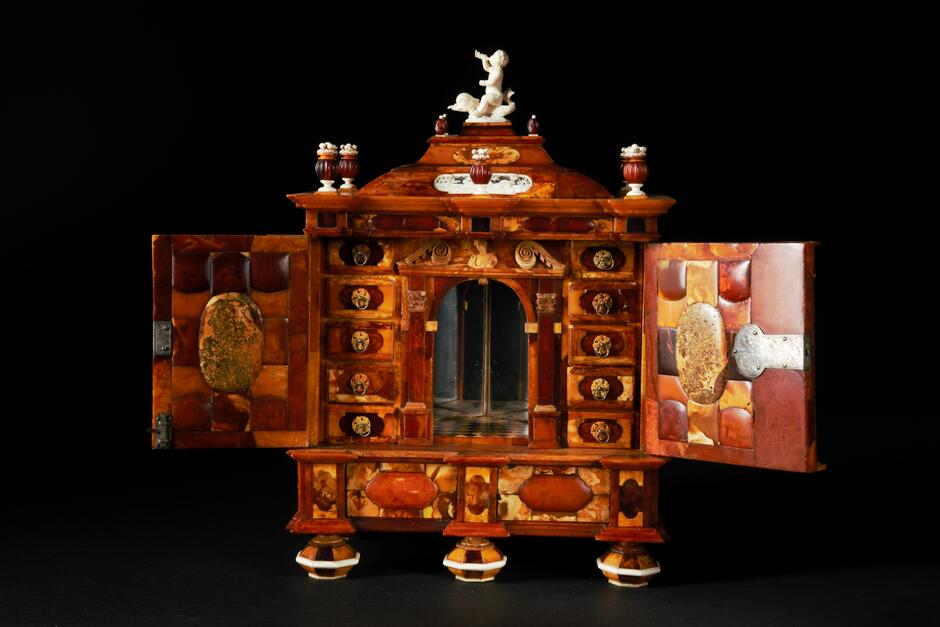 Zdjęcie przedstawia misternie wykonany kabinet z bursztynu, pochodzący z Muzeum Bursztynu. Mebel ma otwarte drzwiczki, ukazując liczne szufladki i ozdobne detale, w tym figurkę na szczycie oraz liczne inkrustacje i ornamenty na frontach.