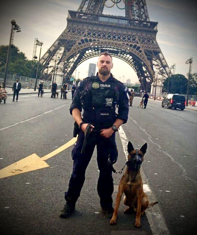 Na zdjęciu widzimy policjanta w mundurze, trzymającego na smyczy psa policyjnego, stojących na tle Wieży Eiffla w Paryżu. Policjant i jego pies stoją na pustej ulicy, a w tle widać kilka osób oraz samochody, co tworzy atmosferę gotowości i bezpieczeństwa.