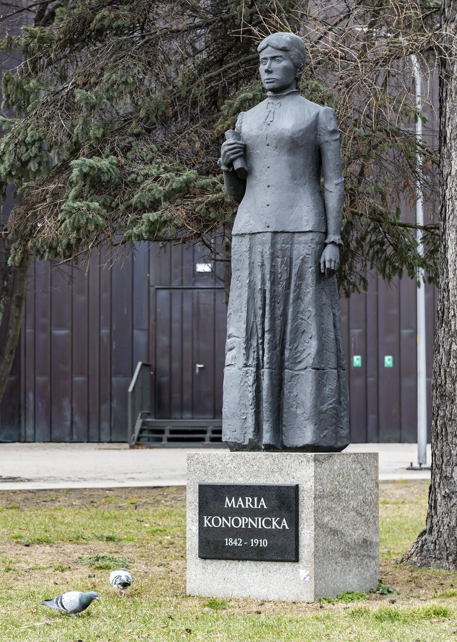 Zdjęcie przedstawia pomnik Marii Konopnickiej, słynnej polskiej pisarki i poetki, znajdujący się na tle drzew. Pomnik wykonany jest z ciemnego materiału i stoi na kamiennym postumencie z inskrypcją MARIA KONOPNICKA 1842-1910 .