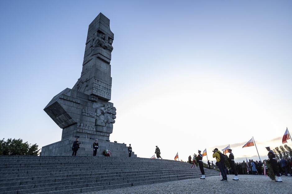 Zdjęcie przedstawia Pomnik Obrońców Wybrzeża na Westerplatte w Gdańsku, widziany z dołu, na tle jasnego nieba. Na schodach przed pomnikiem widoczni są żołnierze w uroczystych mundurach, a także grupa ludzi z flagami, co sugeruje, że odbywają się tam uroczystości upamiętniające.