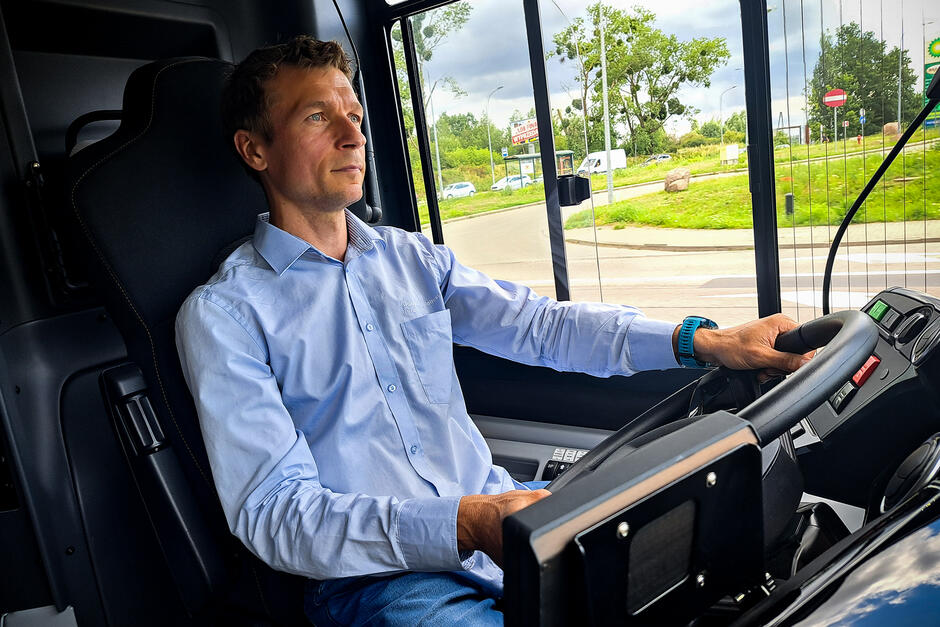 Kierowca autobusu siedzi w kabinie kierowcy i prowadzi autobus. Przez boczne okno pojazdu widać ulice i drzewa.