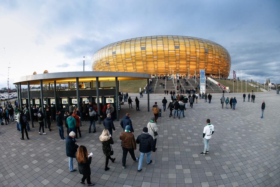 Kibice stoją przed bramkami wejściowymi na mecz. W tle widoczny jest stadion Polsat Plus Arena Gdańsk