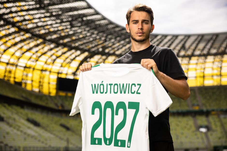 Młody mężczyzna z biało-zieloną koszulką w rękach, na której jest nazwisko Wójtowicz i numer 2027 - bowiem właśnie do 2027 roku zawodnik podpisał kontrakt w Lechią