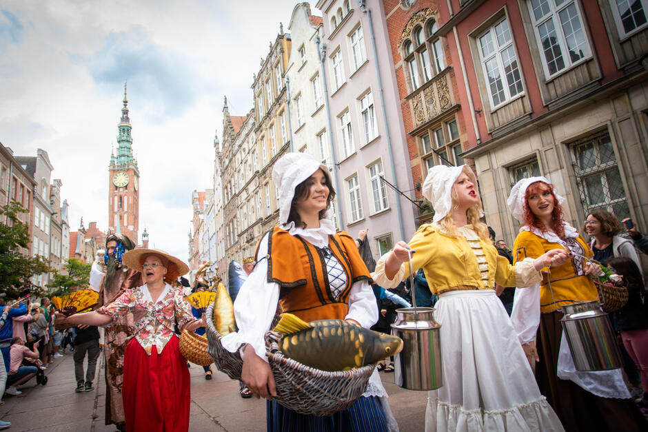 Na zdjęciu widzimy grupę osób ubranych w stroje historyczne, maszerujących przez ulicę w mieście pełnym zabytkowych kamienic. W tle widoczna jest wysoka wieża zegarowa, co sugeruje, że scena rozgrywa się podczas festiwalu lub parady w historycznej części miasta.