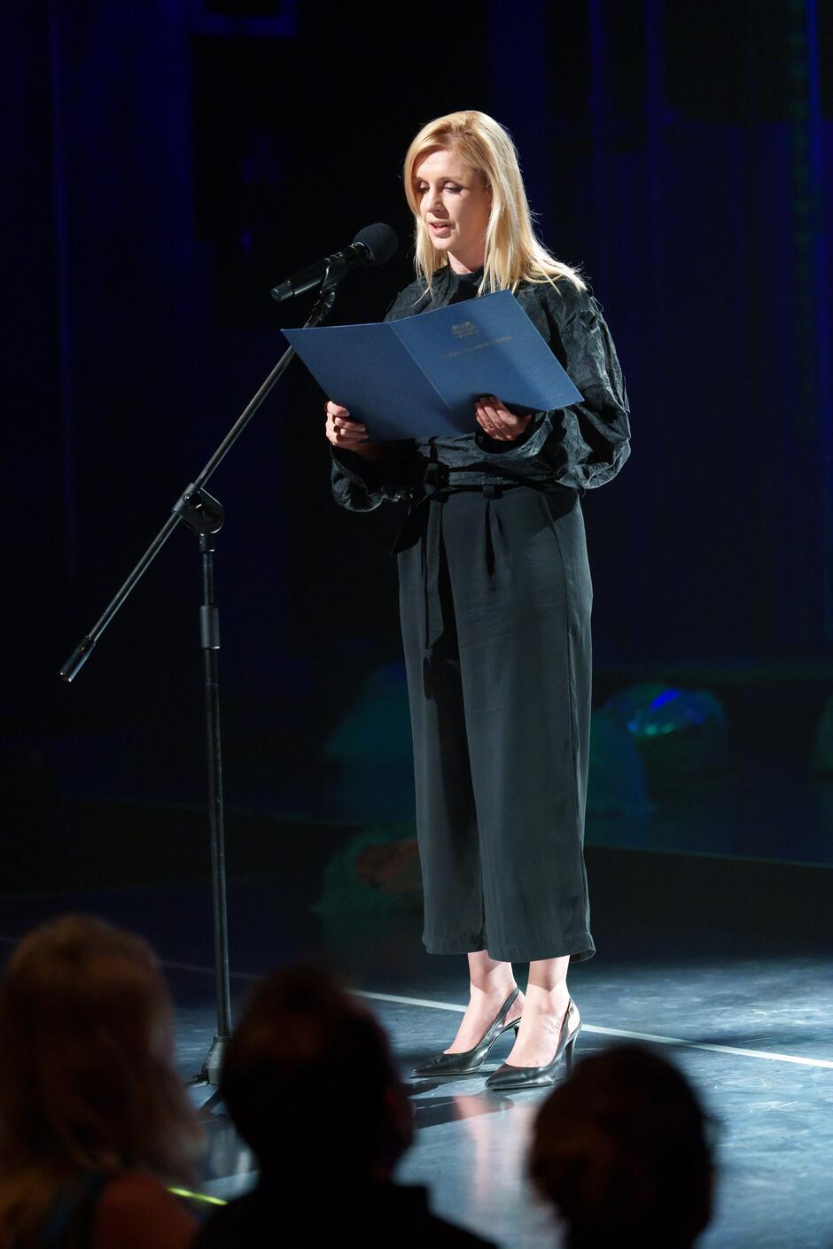 Na zdjęciu widać kobietę stojącą na scenie, trzymającą niebieską teczkę i przemawiającą do mikrofonu. Jest elegancko ubrana w czarne ubranie i buty na obcasie, a w tle widać ciemne oświetlenie sceniczne oraz publiczność siedzącą na widowni