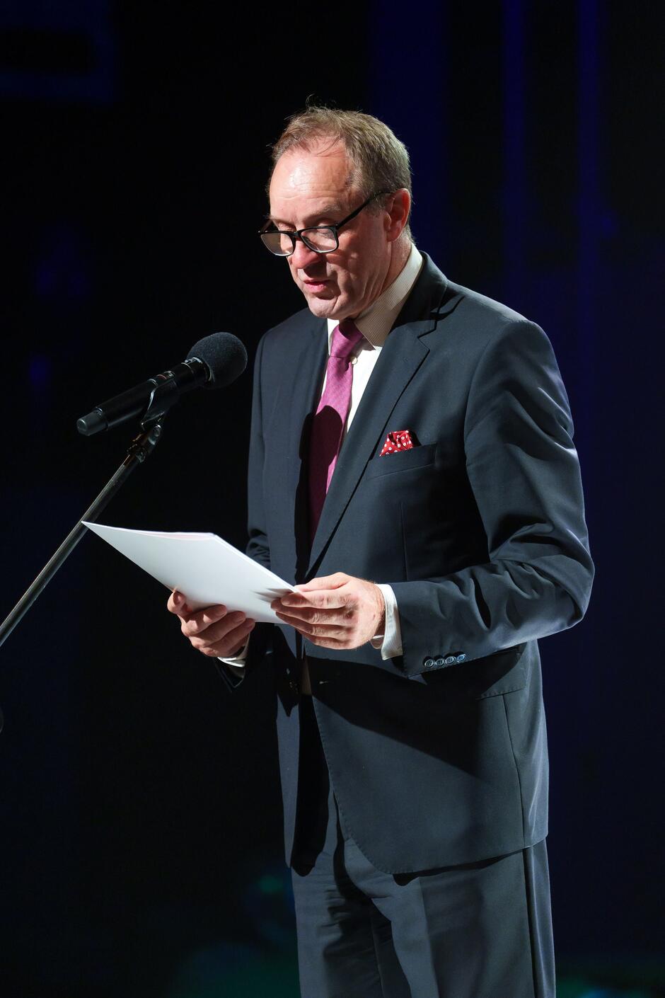 Na zdjęciu widać mężczyznę w eleganckim garniturze, z różowym krawatem i chusteczką w kieszeni, stojącego na scenie i przemawiającego do mikrofonu. Trzyma w rękach kartki z tekstem, a w tle widoczne jest ciemne oświetlenie sceniczne, co sugeruje oficjalne wystąpienie podczas jakiegoś wydarzenia