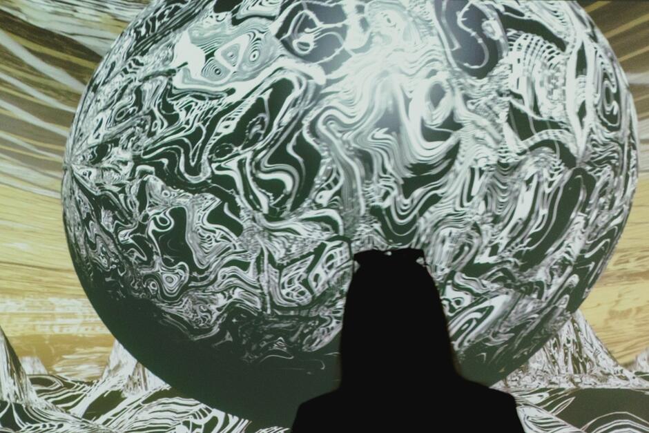 Na zdjęciu widać ciemną sylwetkę osoby stojącej przed dużym, abstrakcyjnie wzorzystym globem. Tło za globem ma faliste, skomplikowane wzory, które dodają całości surrealistycznego charakteru