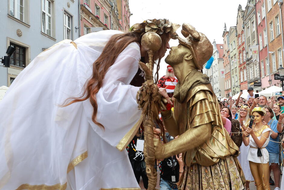 Dwoje uczestników parady, kobieta w wianku i tunice oraz na złoto pomalowany mężczyzna w czapce ze skrzydełkami, charakterystycznej dla Hermesa