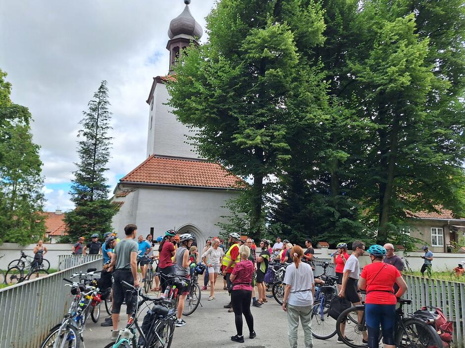 Grupa kilkudziesięciu rowerzystów stoi na moście z rowerami wokół kobiety, przewodniczki, która opowiada im o widocznym w tle kościele.