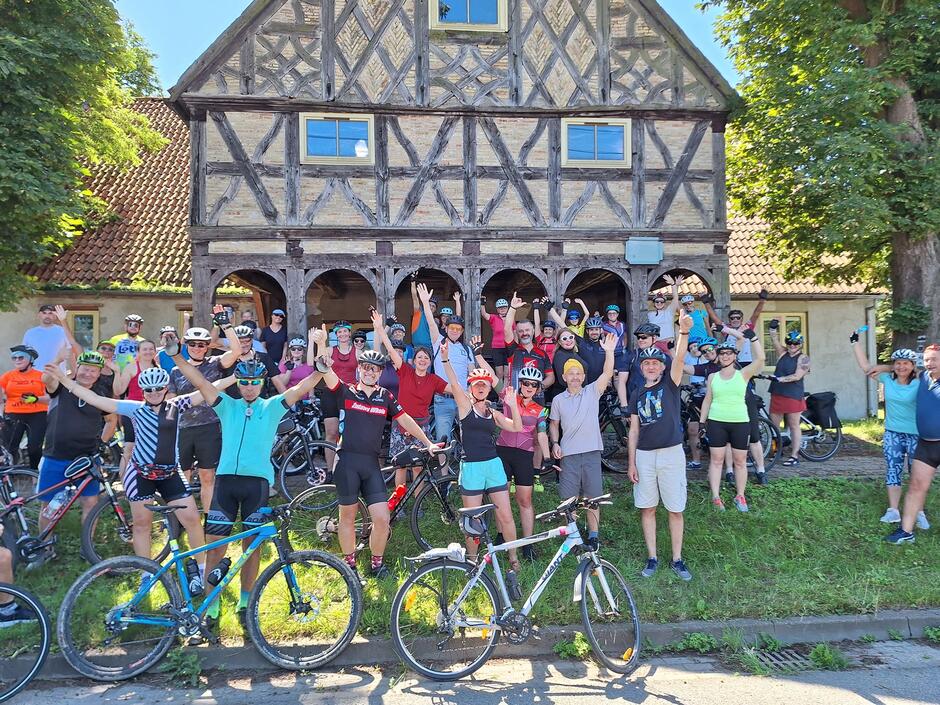 Grupa kilkudziesięciu rowerzystów stoi z rowerami pod zabytkowym domem podcieniowym i macha radośnie do fotografa.