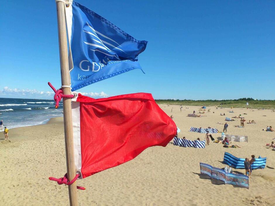 Czerwona flaga powiewa nad plażą, na której plażuje niewielka liczba osób. Na morzy widać wysokie fale.