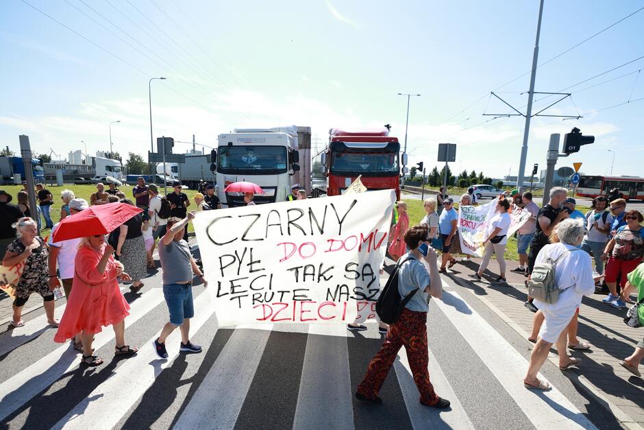 Ludzie z transparentami chodzą po przejściu dla pieszych, za nimi stoją w korku ciężarówki