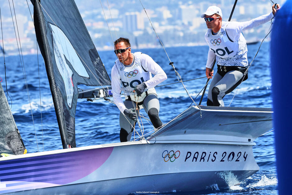 Na zdjęciu widać dwóch żeglarzy w trakcie zawodów olimpijskich na Igrzyskach w Paryżu 2024, na łodzi z napisem "Paris 2024" oraz logiem olimpijskim. Obaj żeglarze są ubrani w białe stroje z napisem "POL" oraz mają na sobie okulary przeciwsłoneczne, a w tle widać rozmyty krajobraz miejski.