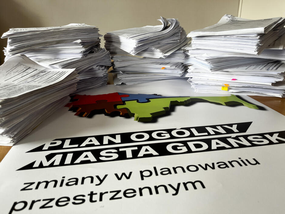 Na zdjęciu widać stosy dokumentów leżące na biurku, obok plakatu z napisem PLAN OGÓLNY MIASTA GDAŃSK, zmiany w planowaniu przestrzennym . Plakat przedstawia również kolorową mapę, co sugeruje, że dokumenty dotyczą zmian w planie zagospodarowania przestrzennego Gdańska.