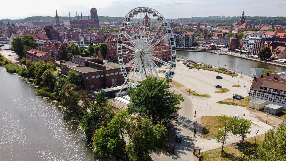 Na zdjęciu widzimy panoramę miasta Gdańska w Polsce, z lotu ptaka. Na pierwszym planie dominuje duży diabelski młyn ustawiony nad brzegiem rzeki Motławy. W tle widoczne są liczne zabytkowe budynki i wieże kościołów, które są charakterystyczne dla starego miasta. Wzdłuż rzeki rozciąga się promenada z kilkoma przechodniami i zaparkowanymi samochodami. Część miasta pokryta jest zielenią, co dodaje uroku temu malowniczemu krajobrazowi. Cała scena jest dobrze oświetlona, co sugeruje, że zdjęcie zostało zrobione w słoneczny dzień. 