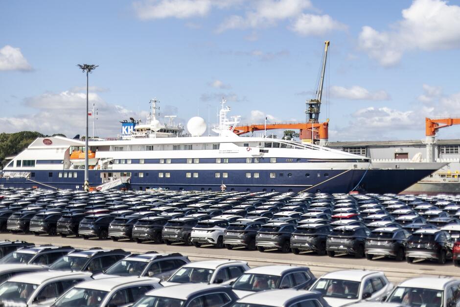 Przy nabrzeżu portowym stoi zacumowany statek wycieczkowy. na pierwszym planie, przed statkiem widać plac, na którym stoją przywiezione do portu samochody. 