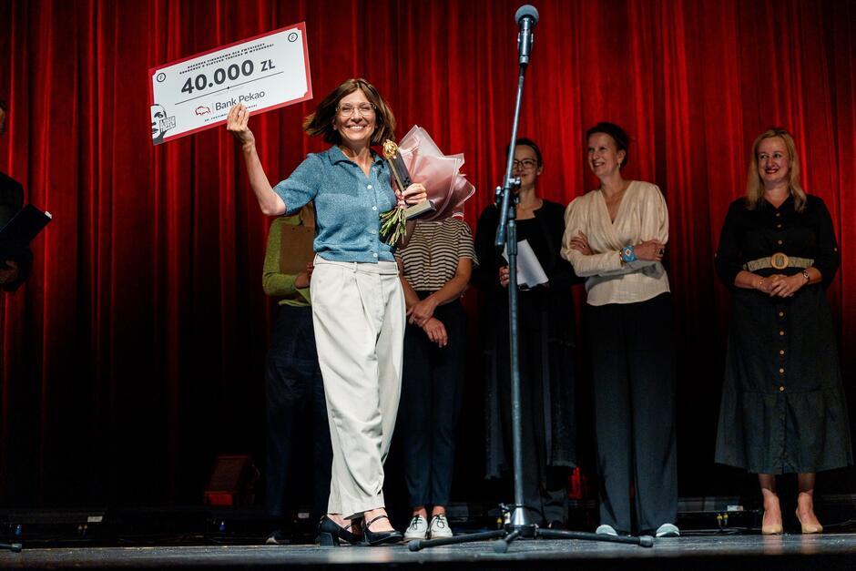 Zdjęcie przedstawia kobietę na scenie teatralnej, trzymającą w rękach symboliczny czek na kwotę 40 000 złotych oraz bukiet kwiatów. W tle za nią stoi grupa osób, które również wydają się brać udział w uroczystości wręczenia nagród, a wszystko dzieje się na tle czerwonej zasłony