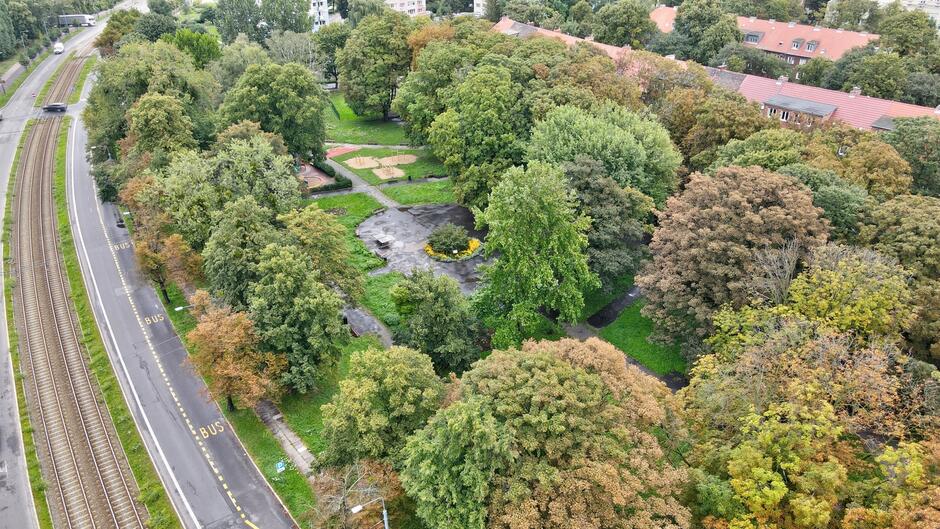 Zdjęcie przedstawia park miejski widziany z lotu ptaka, drzewami i ścieżkami. Po lewej stronie widać tory kolejowe oraz drogę