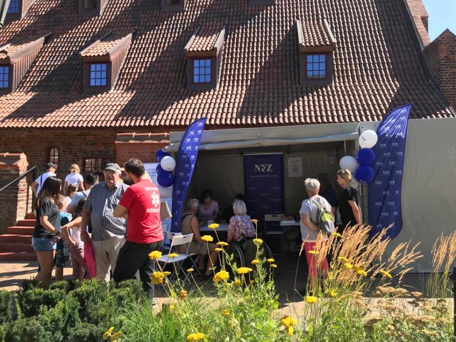 Na tle dachu i ceglanego muru stoisko z niebieskimi flagami z napisem NFZ przed którym stoją ludzie w róznym wieku lekko ubrani 