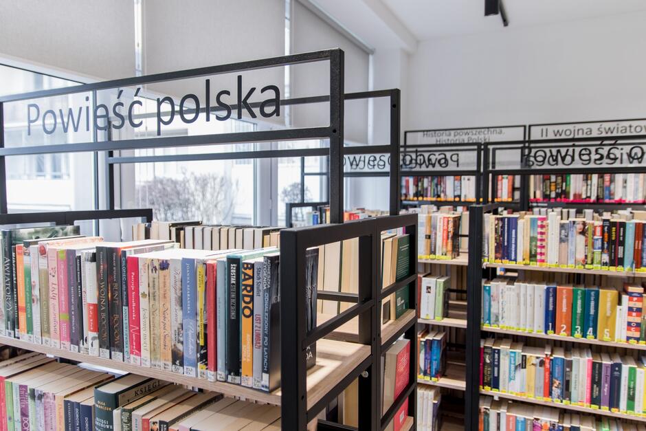 Zdjęcie przedstawia wnętrze biblioteki, gdzie na pierwszym planie widoczne są regały z książkami oznaczone napisem Powieść polska . W tle można dostrzec inne regały z książkami, w tym te z napisami  Historia powszechna  i  II wojna światowa , co sugeruje różnorodność tematyczną dostępnych książek.