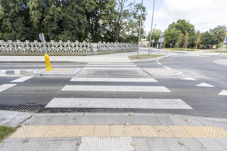 Na zdjęciu widzimy przejście dla pieszych na ulicy, z charakterystycznymi białymi pasami i oznaczeniami dla osób niewidomych na chodniku. W tle znajduje się zielona przestrzeń z drzewami oraz betonowy mur z geometrycznym wzorem.