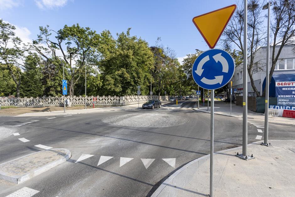 Na zdjęciu widać nowe rondo z wyraźnym oznakowaniem, w tym znakiem drogowym wskazującym ruch okrężny oraz znakiem ustąpienia pierwszeństwa. W tle znajduje się betonowy mur o geometrycznym wzorze oraz otaczająca go zielona przestrzeń z drzewami.