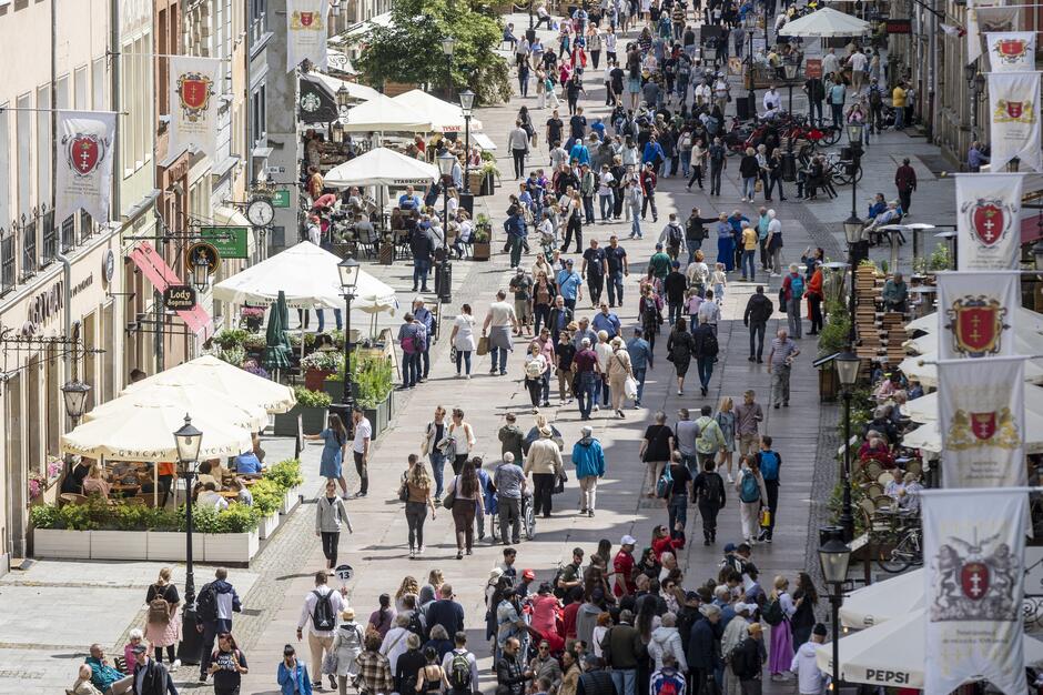 Widok z lotu ptaka na deptak w mieście, którym spacerują tłumy ludzi. Po bokach kadru na fasadach kamienic widać flagi z herbem Gdańska.