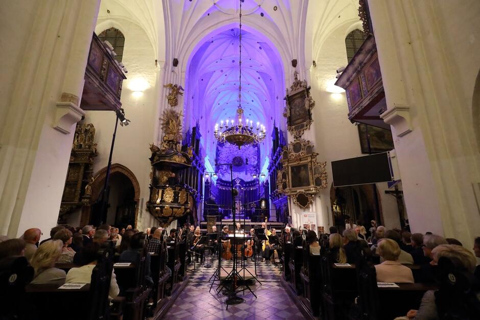 Zdjęcie przedstawia wnętrze kościoła, w którym odbywa się koncert. Widzowie siedzą na ławkach, a na scenie ustawione są instrumenty muzyczne, otoczone bogato zdobionymi elementami architektury kościelnej, z efektownym oświetleniem w kolorze fioletowym