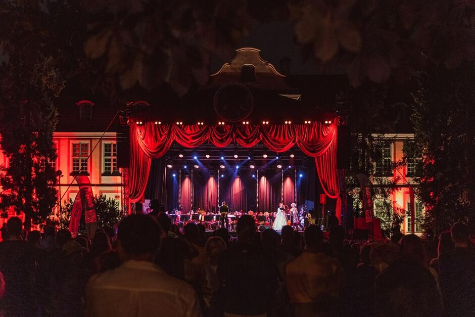 Zdjęcie przedstawia nocny koncert na świeżym powietrzu, zorganizowany przed zabytkowym budynkiem, który jest oświetlony na czerwono. Na scenie ozdobionej czerwoną kurtyną i oświetlonej różnokolorowymi reflektorami występują muzycy i śpiewacy, a przed nimi zgromadziła się licznie publiczność