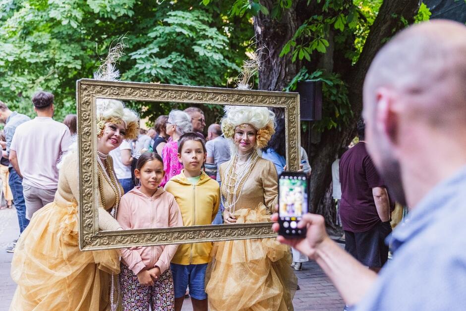 Zdjęcie przedstawia dwie osoby w kostiumach barokowych, pozujące z ramą obrazu razem z dwójką dzieci podczas festiwalu. Mężczyzna robi im zdjęcie telefonem, a w tle widać innych uczestników wydarzenia spacerujących po parku