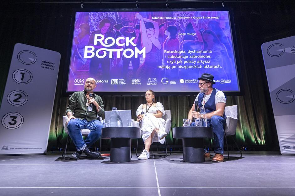 Na zdjęciu widzimy trzech osób siedzących na scenie podczas panelu dyskusyjnego, na tle dużego ekranu z napisem Rock Bottom . W tle znajdują się bannery oraz projekcja przedstawiająca tematykę rotoskopii, dysharmonii i substancji zabronionych w kontekście polskich artystów pracujących nad hiszpańskimi aktorami