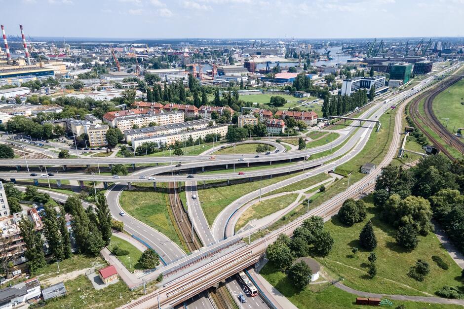 Zdjęcie z lotu ptaka przedstawia rozbudowany węzeł drogowy w miejskiej okolicy, gdzie kilka pasów ruchu i estakad krzyżuje się nad torami kolejowymi. W tle widoczne są zabudowania mieszkalne oraz obszary zielone, co tworzy kontrast między infrastrukturą drogową a miejskim krajobrazem.