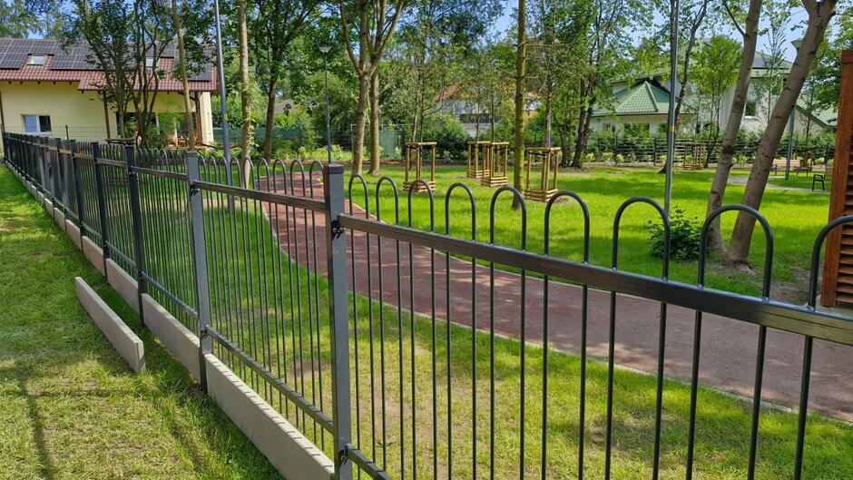 Na zdjęciu widać ogrodzenie oddzielające zadbany park od sąsiednich posesji, w którym znajduje się alejka otoczona trawą oraz młodymi drzewami. W tle widać budynki mieszkalne z panelami słonecznymi na dachach oraz zieloną roślinność otaczającą teren parku.