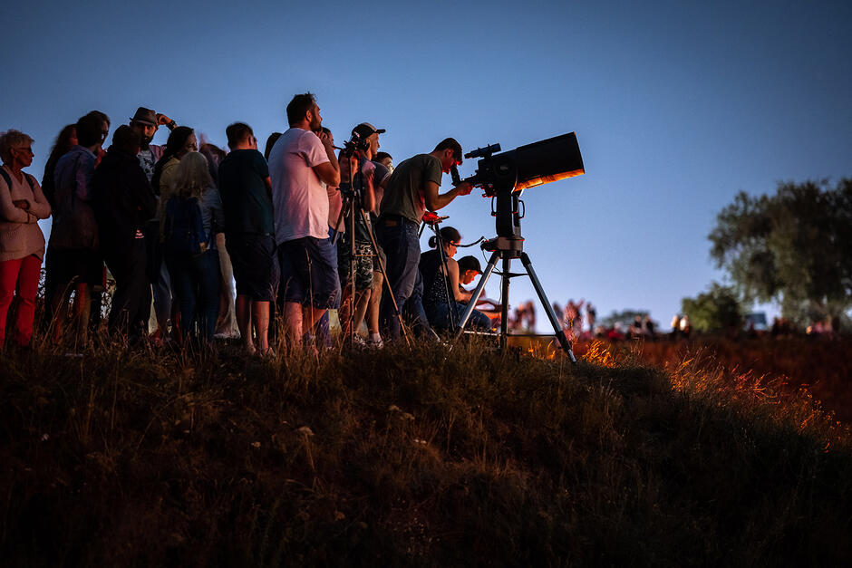 Grupa ludzi stoi późnym wieczorem na obrośniętej trawą górze i obserwuje niebo. Jedna osoba patrzy przez teleskop, inna rozstawiła na statywie aparat fotograficzny.