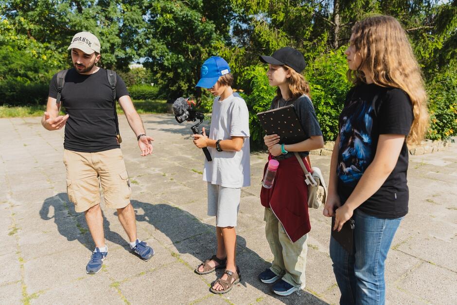 Na zdjęciu widać mężczyznę w beżowej czapce, który prowadzi rozmowę z trzema młodymi osobami stojącymi przed nim, prawdopodobnie uczestnikami warsztatów filmowych. Jedno z dzieci trzyma kamerę na stabilizatorze, a pozostałe mają notatniki, co sugeruje, że są zaangażowani w projekt filmowy lub edukacyjny na świeżym powietrzu.