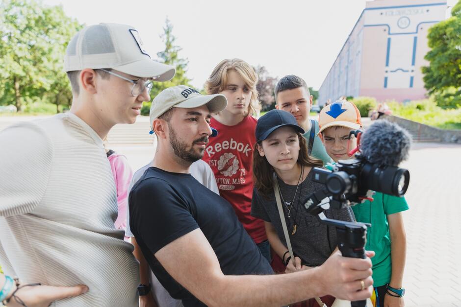 Na zdjęciu widzimy grupę młodych osób, które razem z dorosłym mężczyzną, prawdopodobnie instruktorem, uważnie przyglądają się obrazowi na ekranie kamery umieszczonej na stabilizatorze. Cała grupa jest skupiona i zainteresowana, co sugeruje, że są zaangażowani w proces nauki filmowania lub uczestniczą w warsztatach filmowych na świeżym powietrzu.