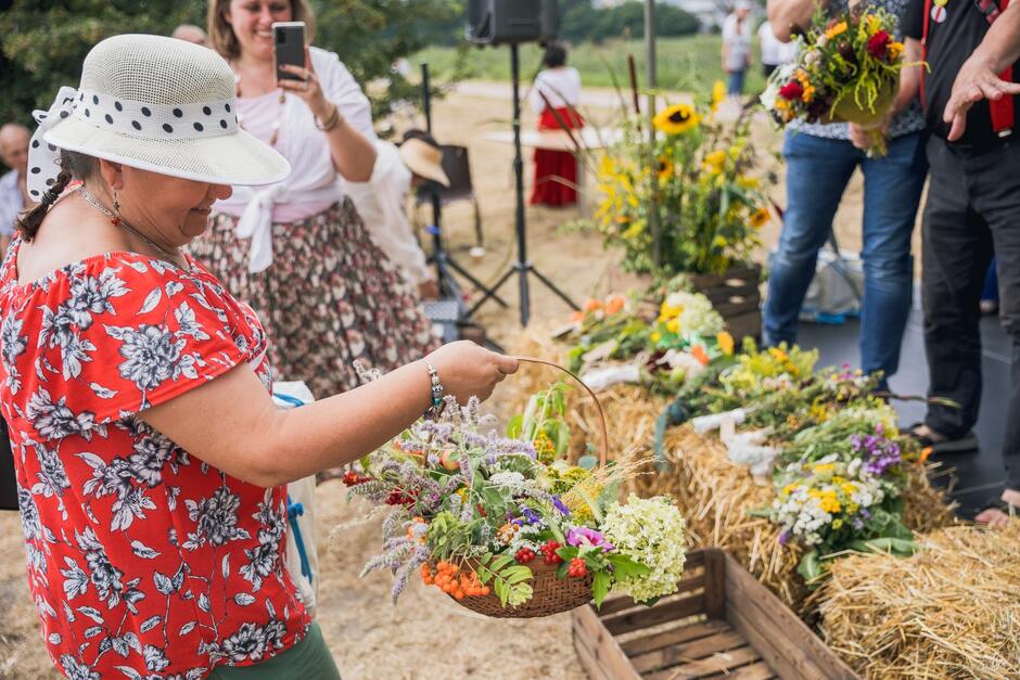 na zdjęciu kobieta w słomkowym kapeluszu trzyma jasny koszyczek wypełniony kwiatami i ziołami z łąki, w tle po lewej widać kobietę która robi tamtej zdjęcie telefonem