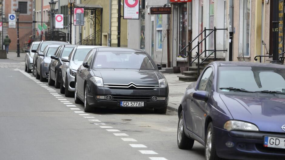 Na zdjęciu widzimy rząd zaparkowanych samochodów wzdłuż ulicy, która znajduje się w mieście. Samochody stoją po prawej stronie jezdni, zaparkowane równolegle do krawężnika. Na pierwszym planie widoczny jest ciemnoszary samochód marki Citroën, z widoczną tablicą rejestracyjną z numerem zaczynającym się od GD , co sugeruje, że zdjęcie zostało zrobione w Gdańsku, w Polsce. W tle widać różnokolorowe budynki z witrynami sklepów oraz chodnik, na którym nie ma zbyt wielu ludzi. Po lewej stronie ulicy widoczne są również latarnie oraz znaki drogowe. Pogoda wydaje się być pochmurna, a ulica jest spokojna i niezatłoczona.       Na zdjęciu widzimy rząd zaparkowanych samochodów wzdłuż ulicy, która znajduje się w mieście. Samochody stoją po prawej stronie jezdni, zaparkowane równolegle do krawężnika. Na pierwszym planie widoczny jest ciemnoszary samochód marki Citroën, z widoczną tablicą rejestracyjną z numerem zaczynającym się od  GD , co sugeruje, że zdjęcie zostało zrobione w Gdańsku, w Polsce. W tle widać różnokolorowe budynki z witrynami sklepów oraz chodnik, na którym nie ma zbyt wielu ludzi. Po lewej stronie ulicy widoczne są również latarnie oraz znaki drogowe. Pogoda wydaje się być pochmurna, a ulica jest spokojna i niezatłoczona.       