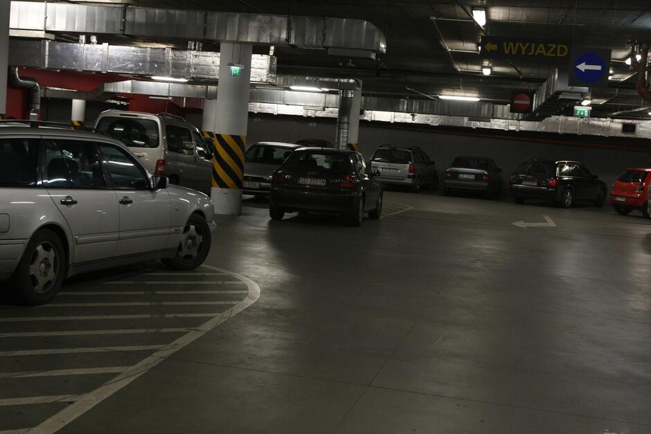 Na zdjęciu widać podziemny parking z zaparkowanymi samochodami. Wnętrze parkingu jest oświetlone sztucznym światłem, a ściany oraz filary są pokryte szaro-żółtymi pasami ostrzegawczymi. Kilka pojazdów jest zaparkowanych w różnych miejscach, a na jednym z filarów widnieje znak informujący o kierunku wyjazdu ( WYJAZD ) w kolorze żółtym z czarną strzałką. Na podłodze widać oznaczenia poziome, które wskazują miejsca parkingowe oraz kierunek ruchu. Cała scena jest typowa dla podziemnych parkingów w większych centrach handlowych lub biurowcach, gdzie panuje spokojna i uporządkowana atmosfera.